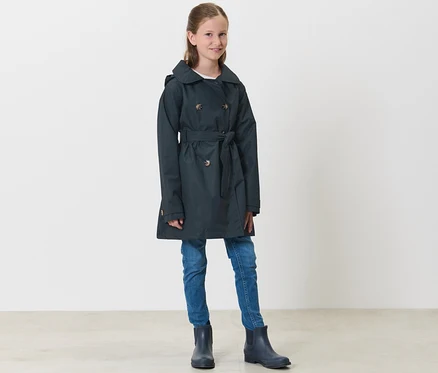Une fille debout porte un trench-coat tout-temps marine pour enfant, un jean et des bottes en caoutchouc.