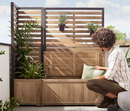 Une femme place un coussin sur un module latéral de paravent «Elin» avec banc de rangement. À l'arrière-plan se trouve un module d'angle de paravent «Elin» avec jardinière.