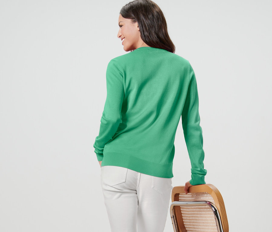 Une femme regarde par-dessus son épaule et porte un cardigan vert en maille fine et un pantalon blanc.