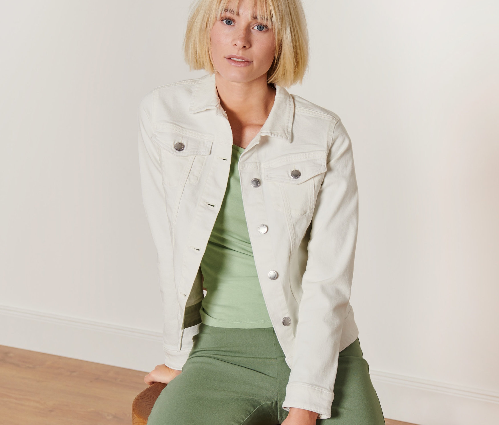 Une femme blonde est assise sur un tabouret et porte une veste en jean de couleur crème.