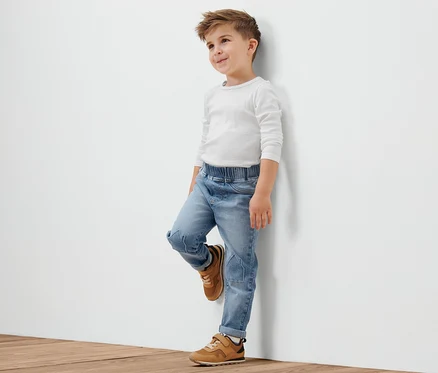 Un garçon portant un t-shirt blanc et un denim élastique pour tout-petit, avec broderie motif petits maisons est appuyé contre un mur blanc.