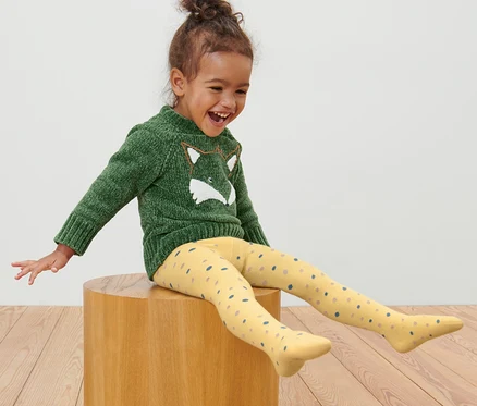 Une fille est assise sur un tabouret en bois et porte 5 collants pour tout-petits.