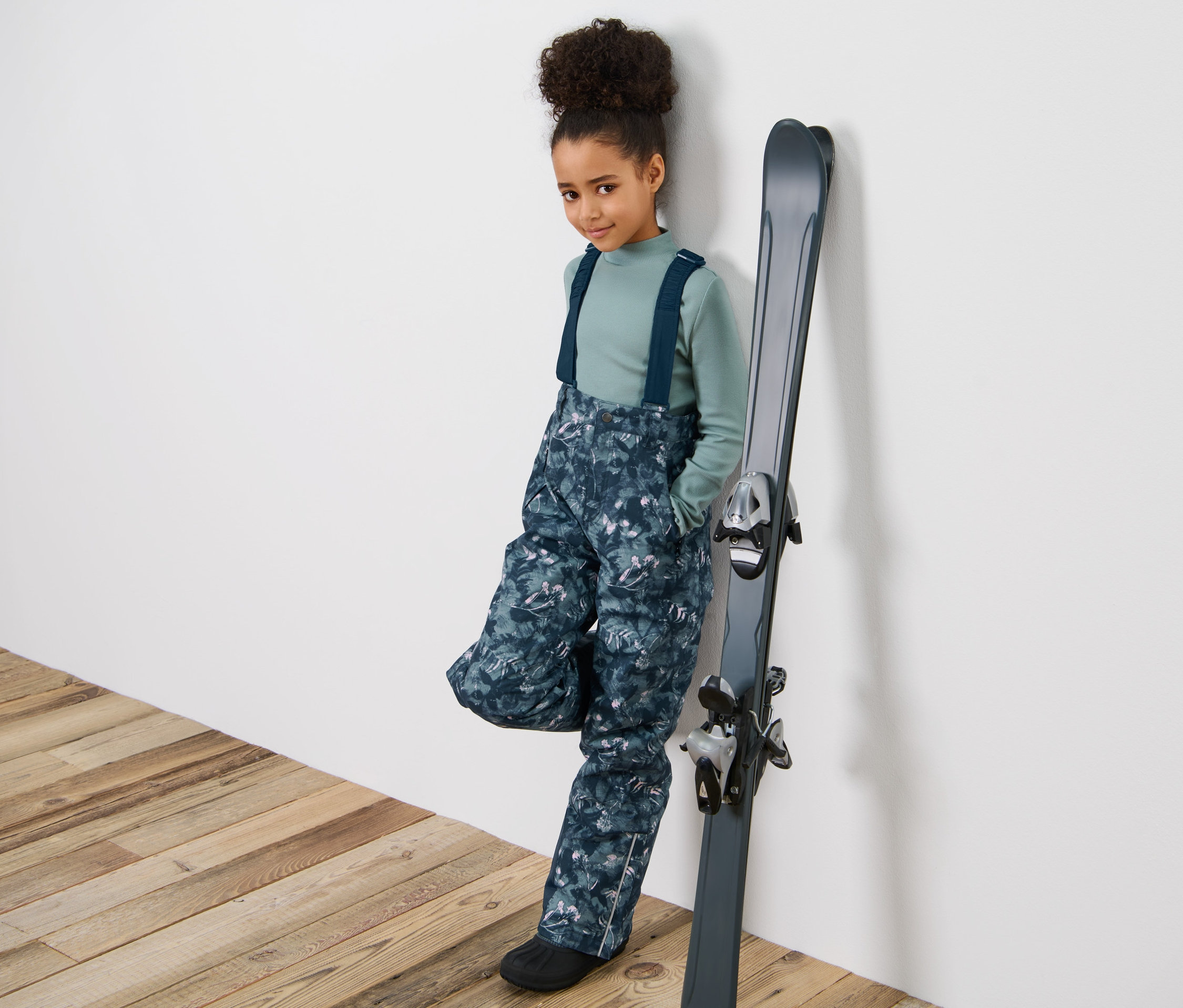 Fille portant un pantalon de ski pour enfant avec imprimé intégral, appuyée contre des skis.