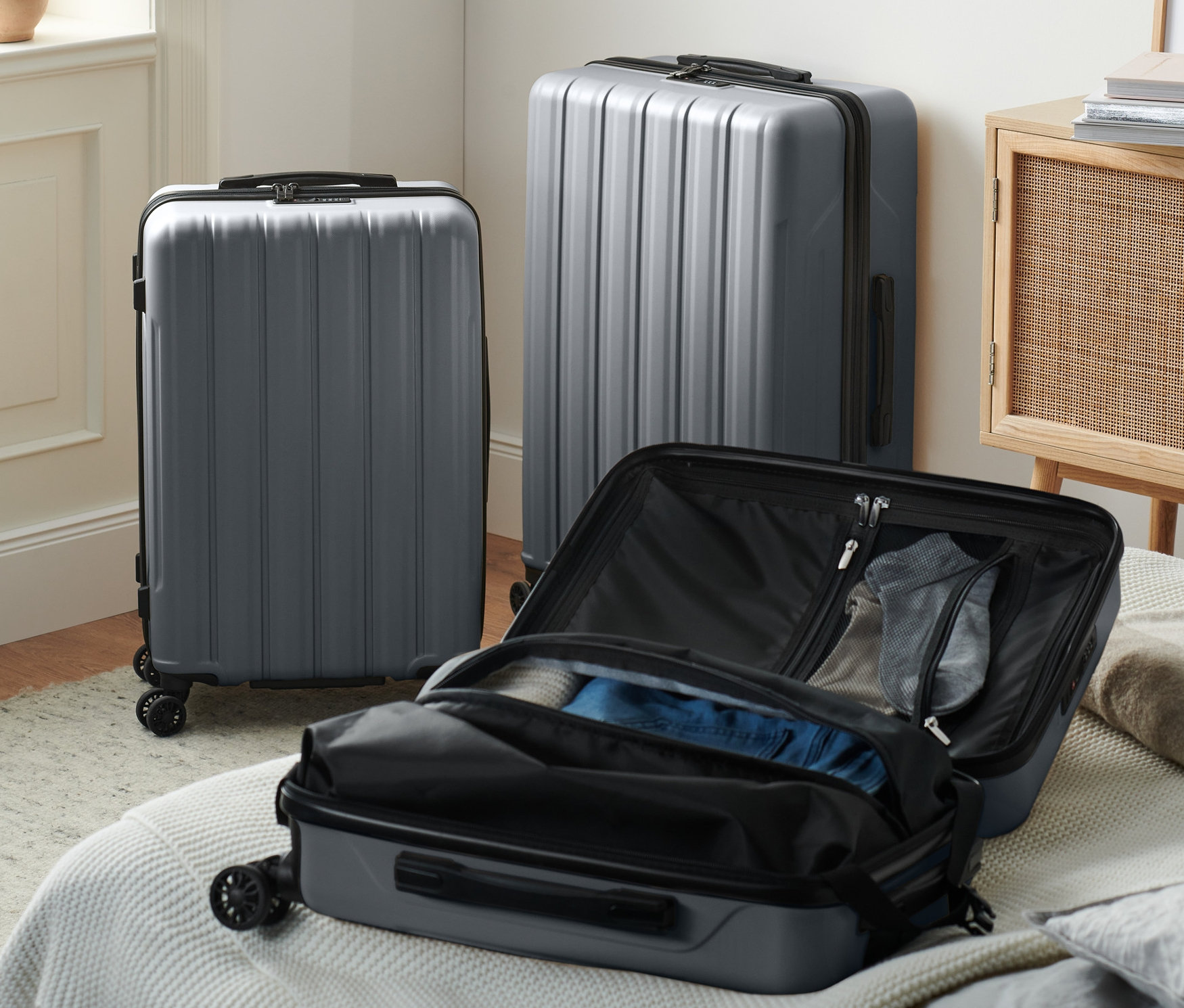 Trois valises rigides grises (petite, moyenne et grande) sont posées dans une pièce, dont une ouverte sur un lit.