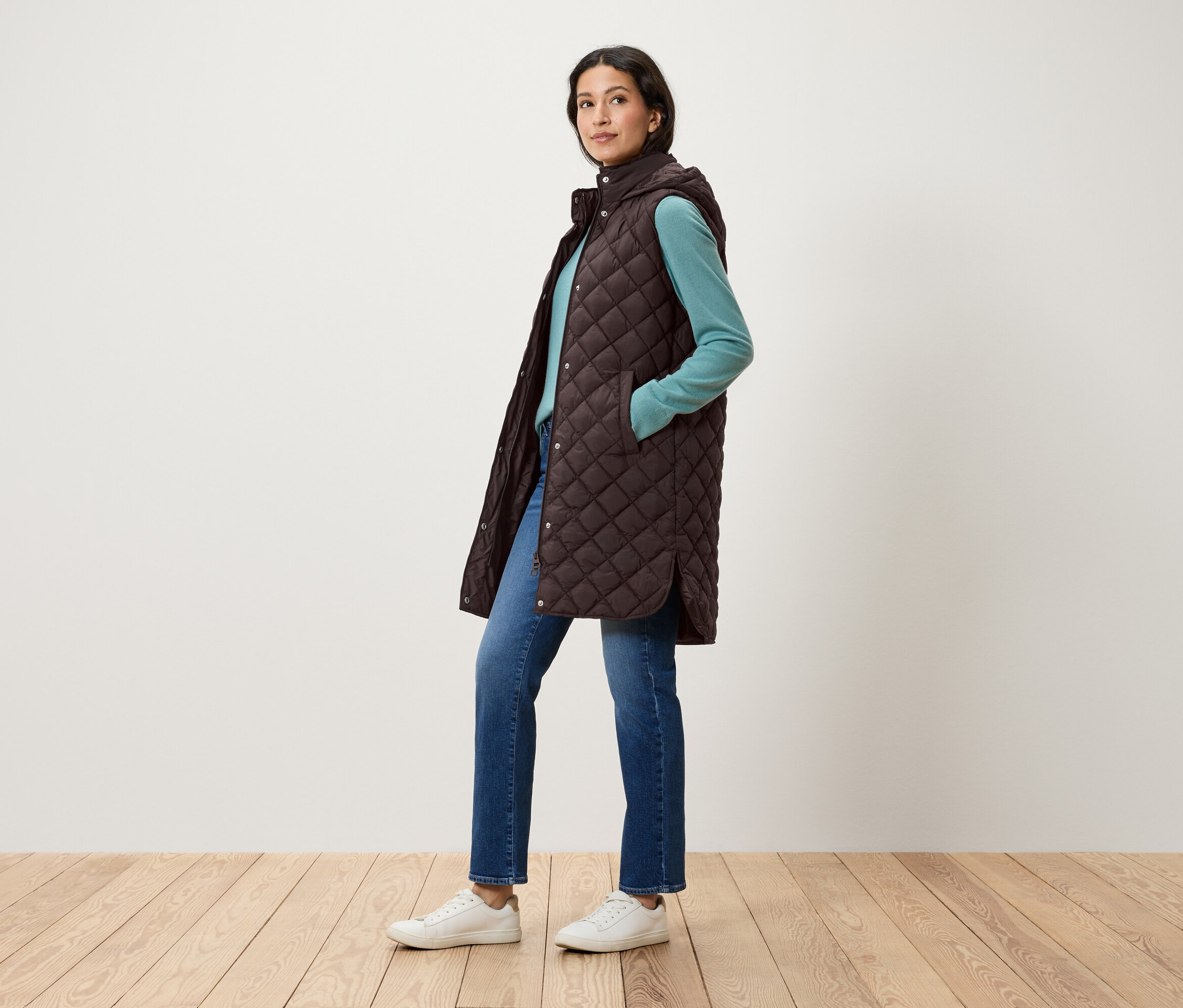 Femme posant dans un gilet matelassé marron à capuche, un pull bleu, un jean et des chaussures blanches.