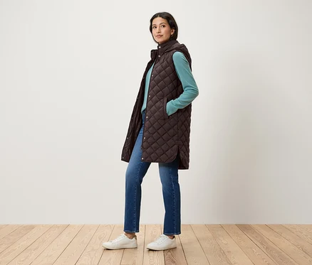 Femme posant dans un gilet matelassé marron à capuche, un pull bleu, un jean et des chaussures blanches.