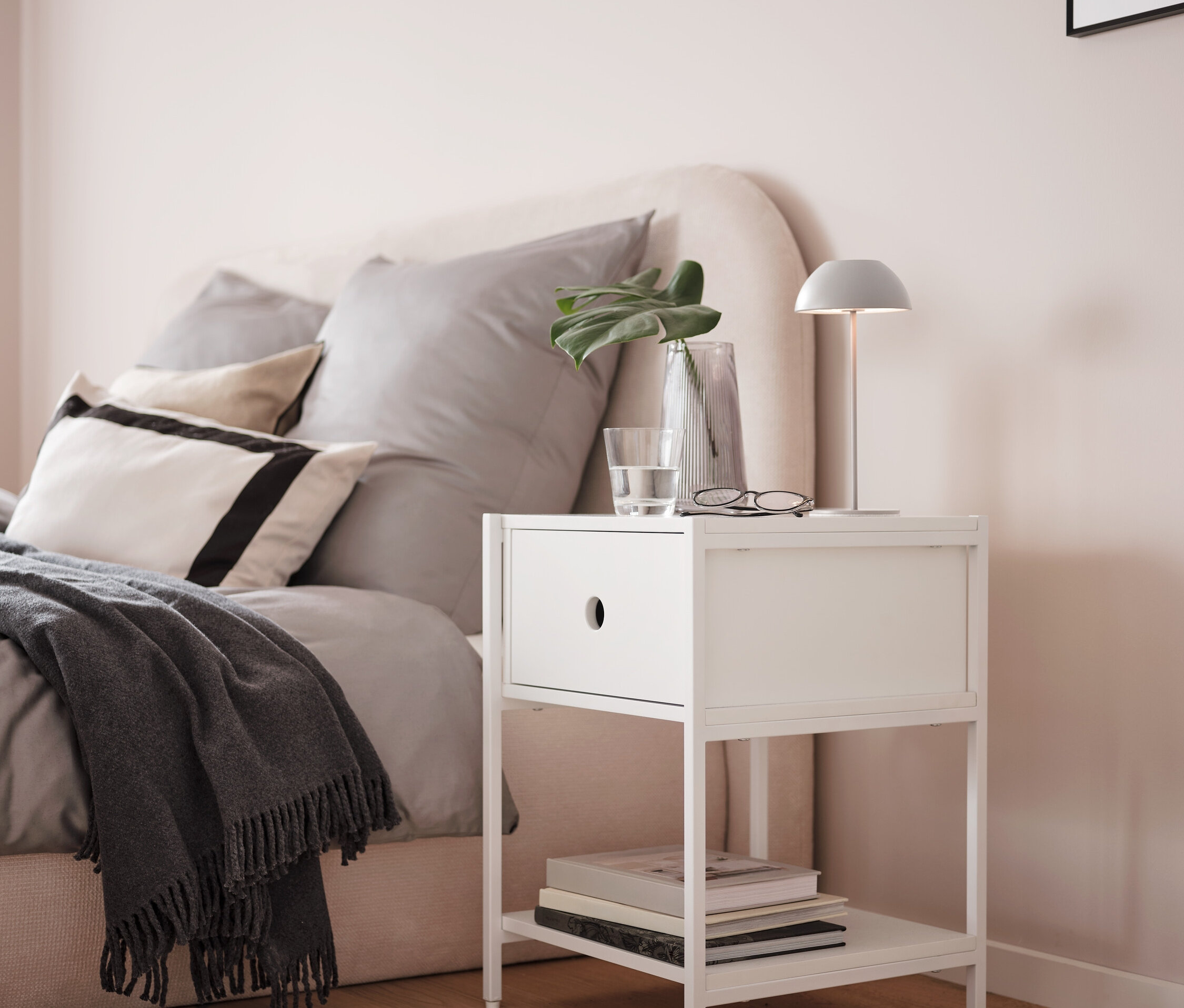 Une lampe de table rechargeable à LED grise est posée sur une table de chevet blanche à côté d'un lit.