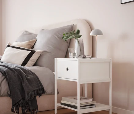 Une lampe de table rechargeable à LED grise est posée sur une table de chevet blanche à côté d'un lit.