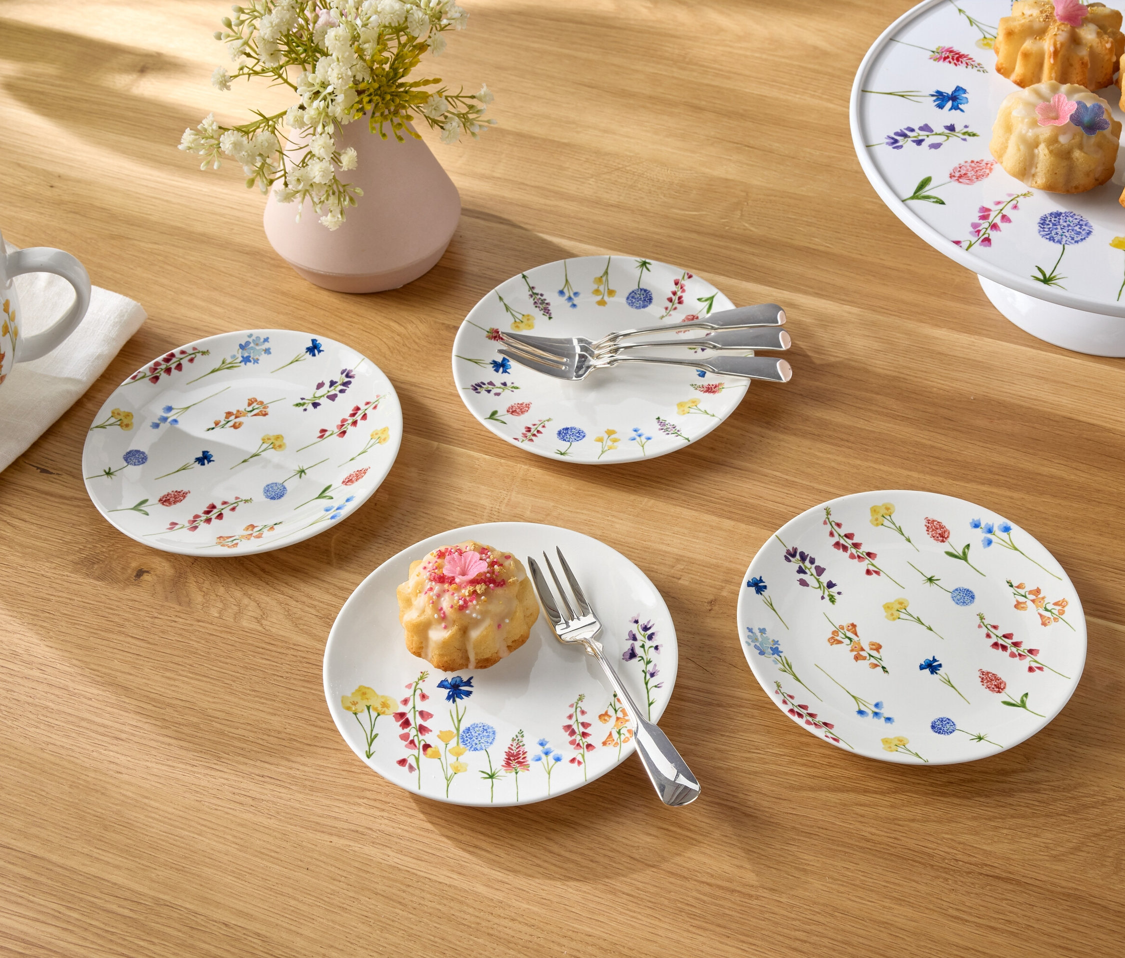 Sur une table en bois, il y a une tasse de café, un vase de fleurs, des assiettes et des petits gâteaux.