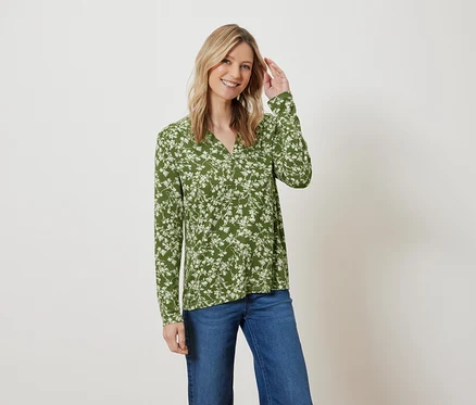 Femme blonde posant dans un chemisier vert à fleurs et un jean bleu.