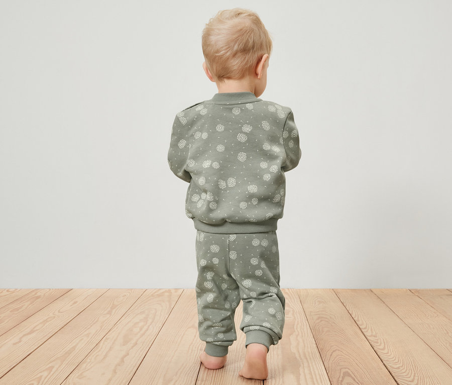 Un jeune enfant vu de dos marche pieds nus sur un plancher en bois et porte un survêtement vert.