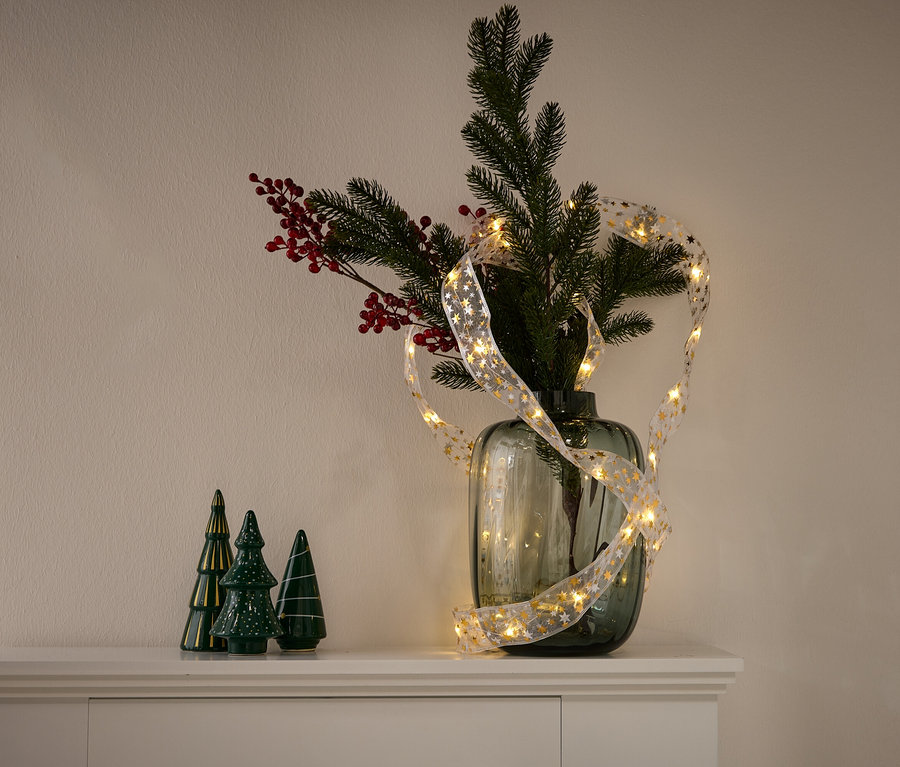 Un vase avec des branches de conifères, des baies rouges et une guirlande lumineuse avec des étoiles dorées. Trois sapins en céramique verte se trouvent à gauche.
