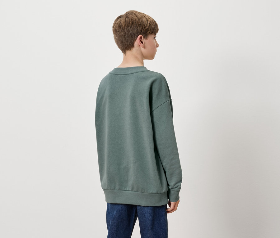 De dos, un garçon aux cheveux bruns courts, portant un sweat-shirt vert et un jean bleu.