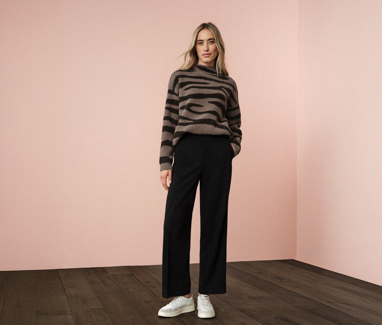 Une femme se tient devant un mur rose portant un pull jacquard Street One, un pantalon noir et des baskets blanches.