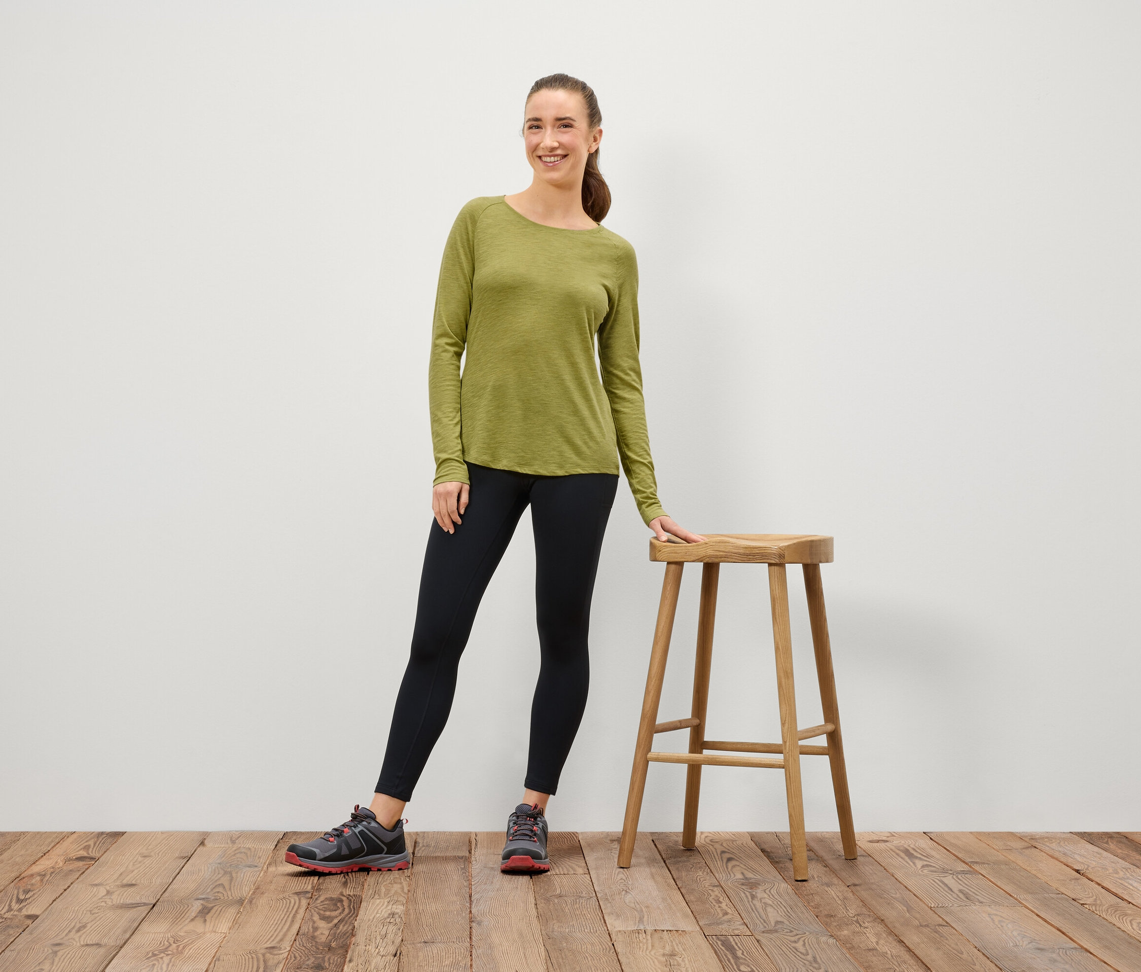 Une femme portant un t-shirt à manches longues vert olive et un legging noir se tient à côté d'un tabouret en bois.