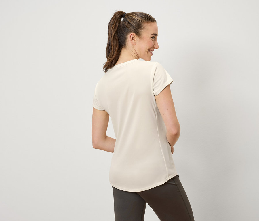 Une femme aux cheveux bruns attachés en queue de cheval sourit et regarde vers la gauche. Elle porte un t-shirt crème et un pantalon marron.