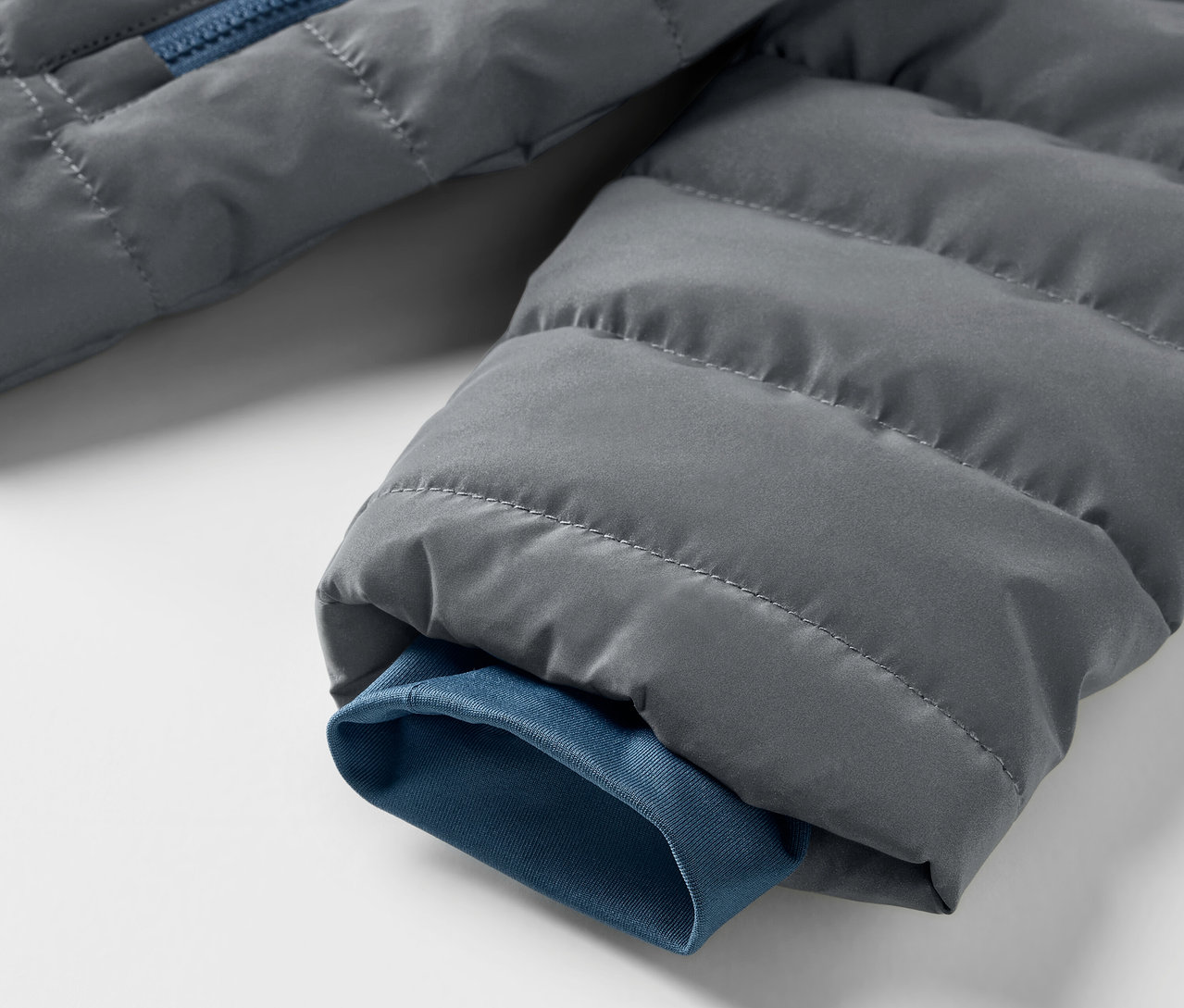 Gros plan sur une veste matelassée grise avec une fermeture éclair bleue et un bord de manche. Veste matelassée réfléchissante pour enfant.