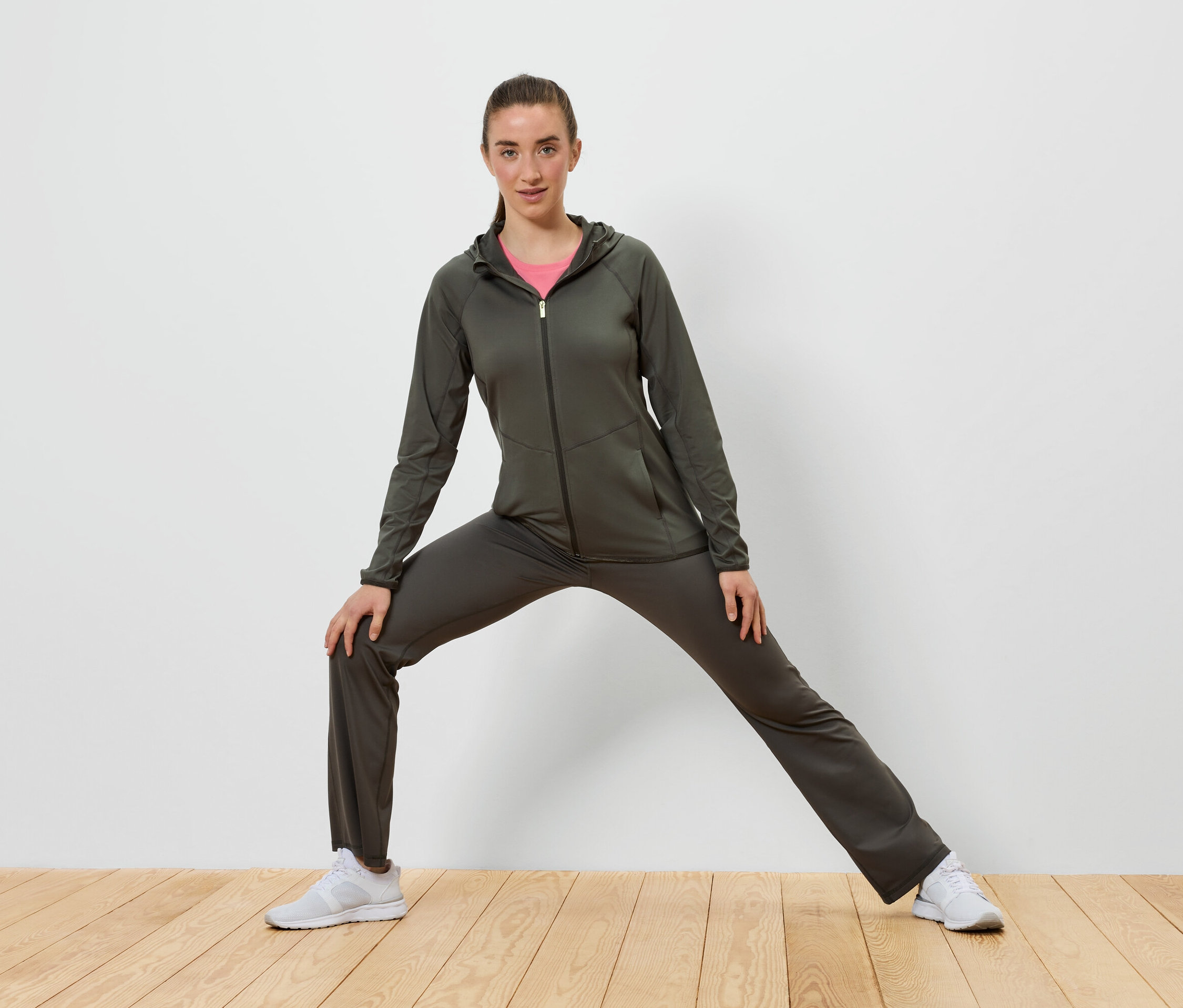 Femme en survêtement vert olive s'étirant sur un plancher en bois devant un mur blanc.