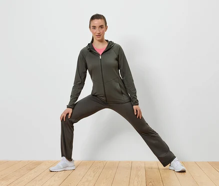 Femme en survêtement vert olive s'étirant sur un plancher en bois devant un mur blanc.