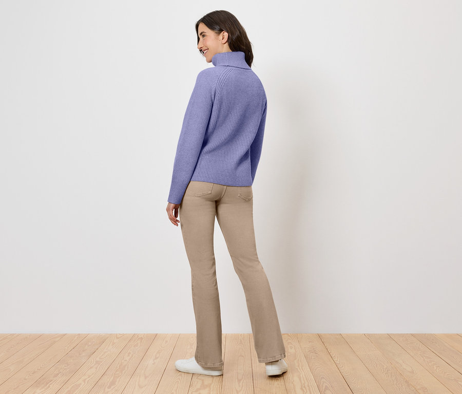 Une femme debout, vue de dos, portant un pull violet à col roulé et un pantalon beige.