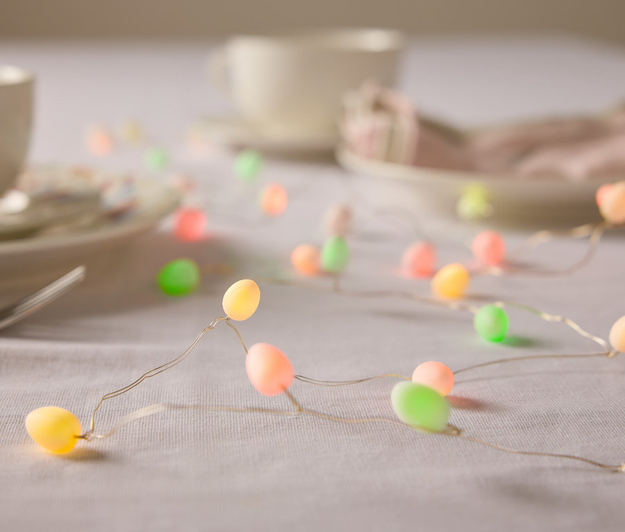 Gros plan d'une table avec de la vaisselle et une guirlande lumineuse en forme d'œufs de Pâques aux couleurs pastel.