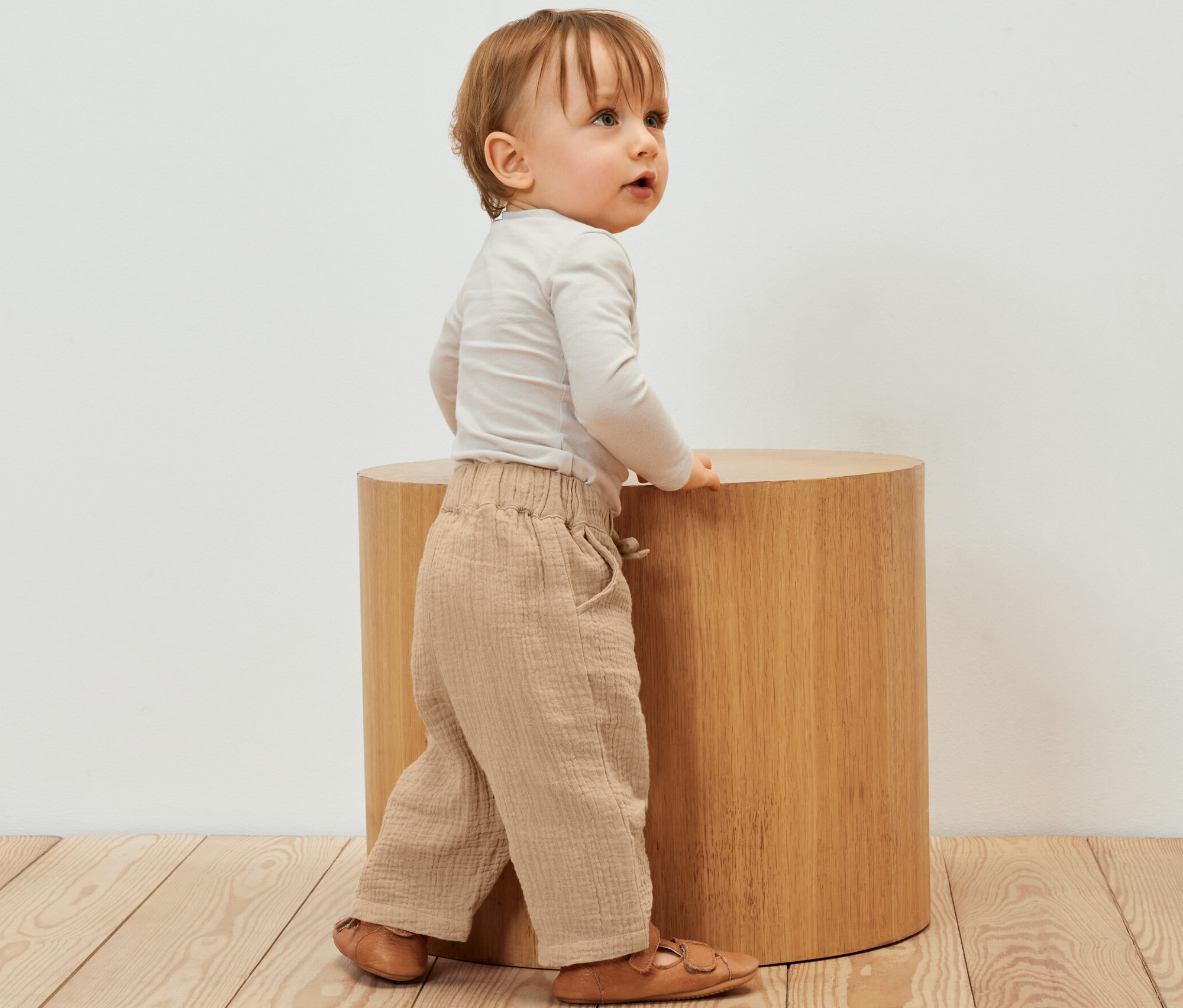 Un enfant se tient à côté d'un tabouret en bois et porte un pantalon en mousseline pour enfant.