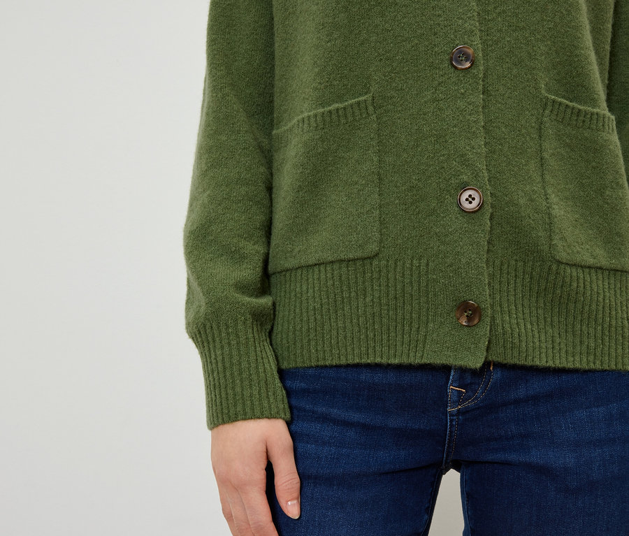 Gros plan d'une femme portant un cardigan olive avec des boutons marron et des poches, associé à un jean bleu foncé.
