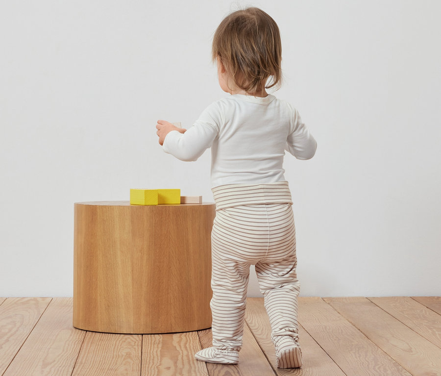 Bébé debout, dos tourné, jouant avec des jouets à côté de 3 grenouillères pour bébé.