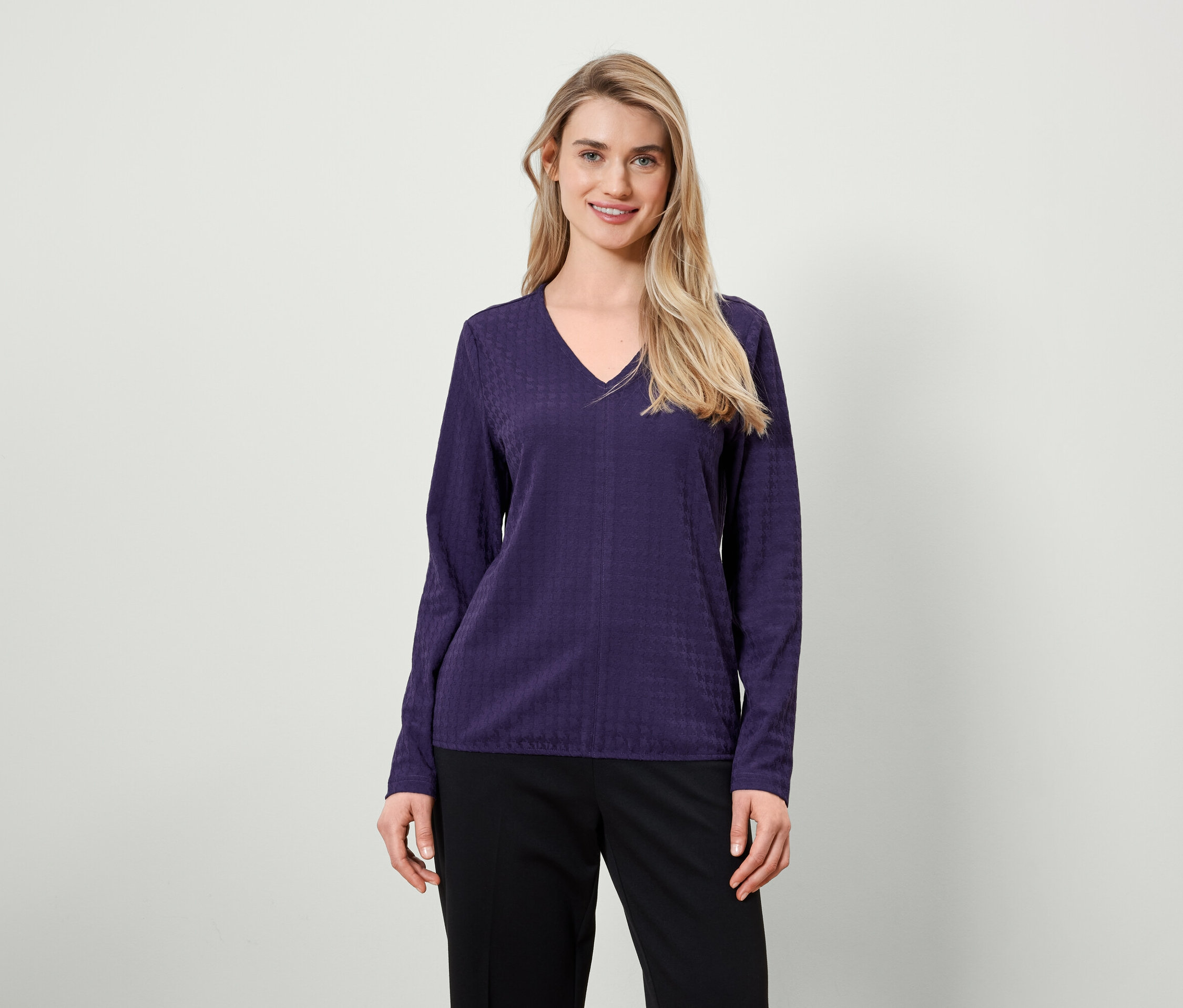 Blonde aux cheveux longs posant dans un chemisier violet à manches longues et un pantalon noir.