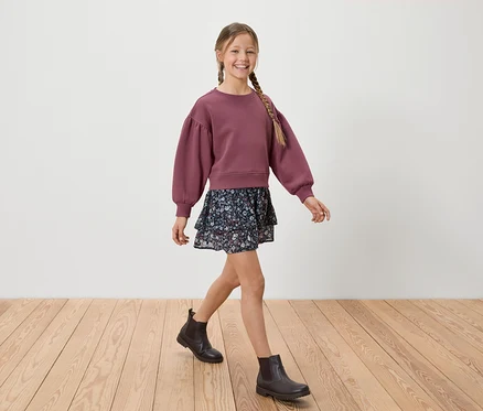 Une jeune fille souriante avec une tresse marche sur un plancher en bois, portant un sweat-shirt bordeaux, une jupe fleurie et des bottes noires.