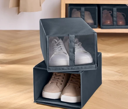 Deux boîtes à chaussures en tissu gris sont empilées l'une sur l'autre. Des baskets blanches sont dans la boîte supérieure et des baskets marron dans la boîte inférieure. Une étagère en bois avec des chaussures est en arrière-plan.