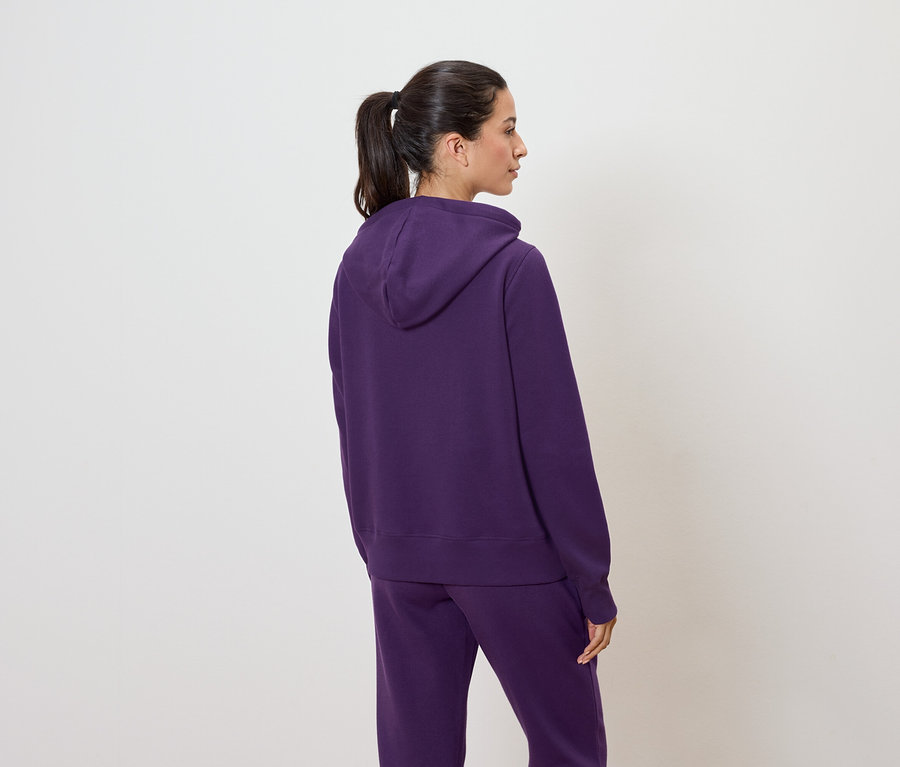Une femme, dos à la caméra, porte un sweat à capuche violet et un pantalon de jogging.