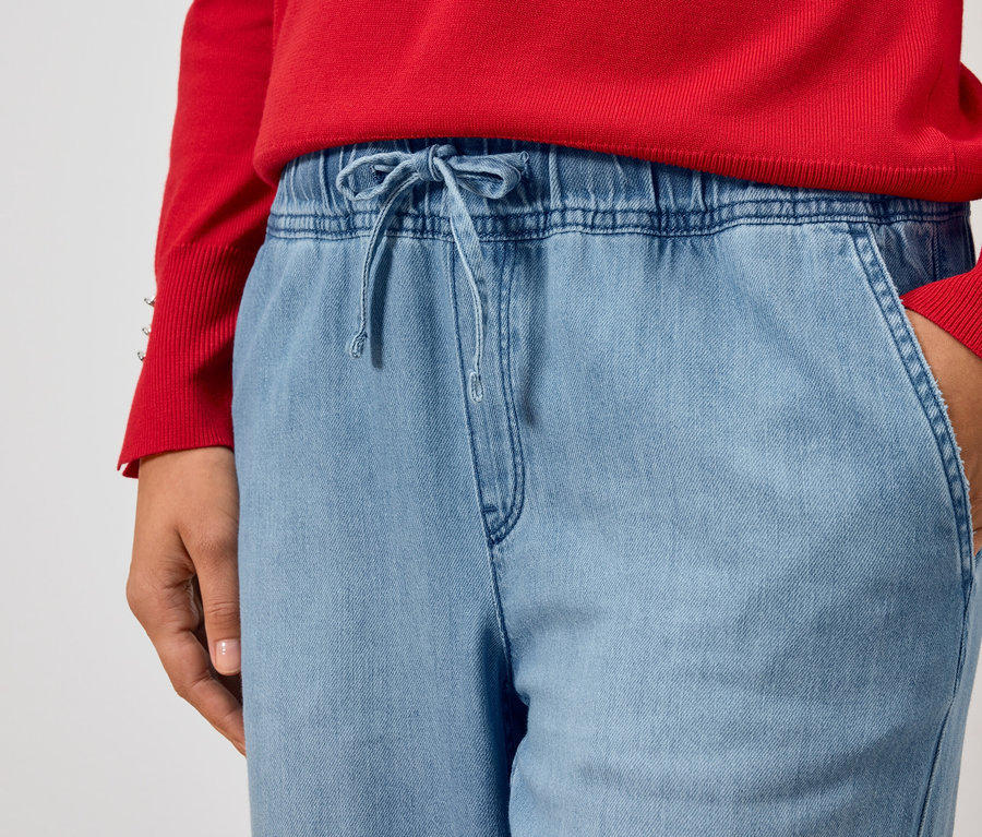 Vue partielle d'une personne portant un pull rouge et un jean bleu clair, la main dans la poche.