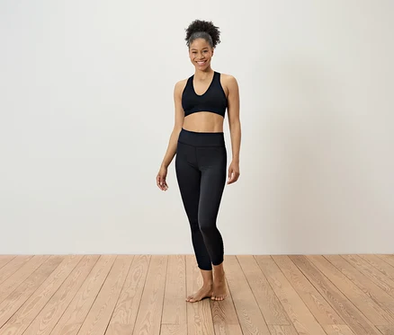 Une femme en tenue de sport noire se tient sur un plancher en bois devant un mur blanc.