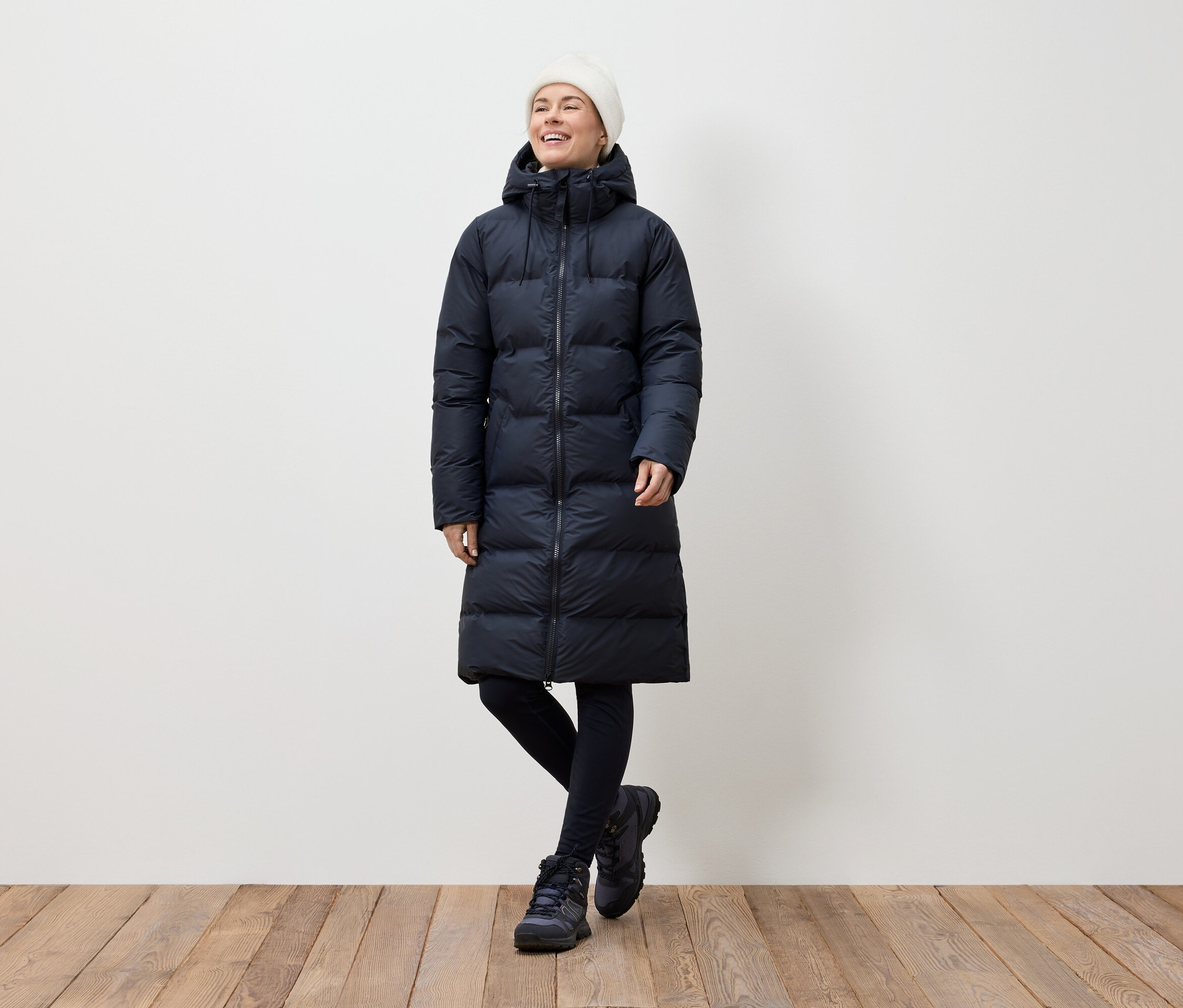 Une femme portant un long manteau en duvet bleu marine, des leggings noirs et des bottes d'hiver se tient sur un plancher en bois et sourit.