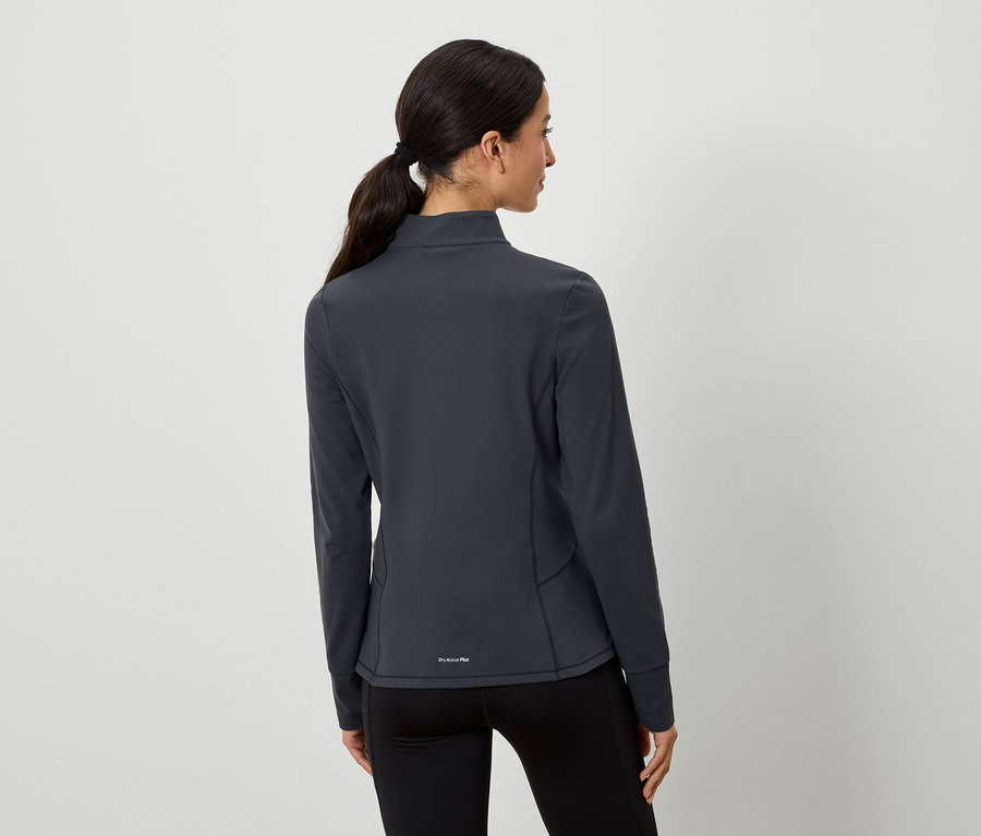 Une femme aux cheveux bruns attachés en queue de cheval pose dos à l'appareil photo. Elle porte une veste de sport grise et un legging noir.