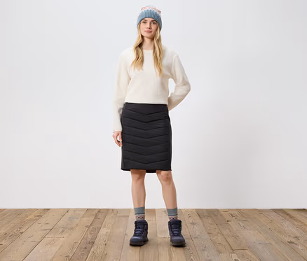 Une femme se tient sur un plancher en bois et porte un pull crème, une jupe noire, des bottes et un bonnet tricoté.