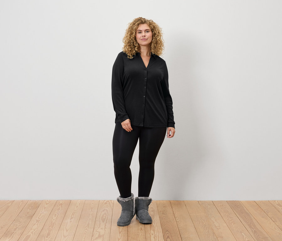 Une femme aux cheveux blonds se tient debout et porte un legging noir, une chemise noire à manches longues et des bottes grises.
