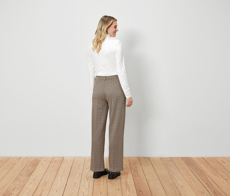 Une femme pose dans un haut blanc et un pantalon à carreaux marron.