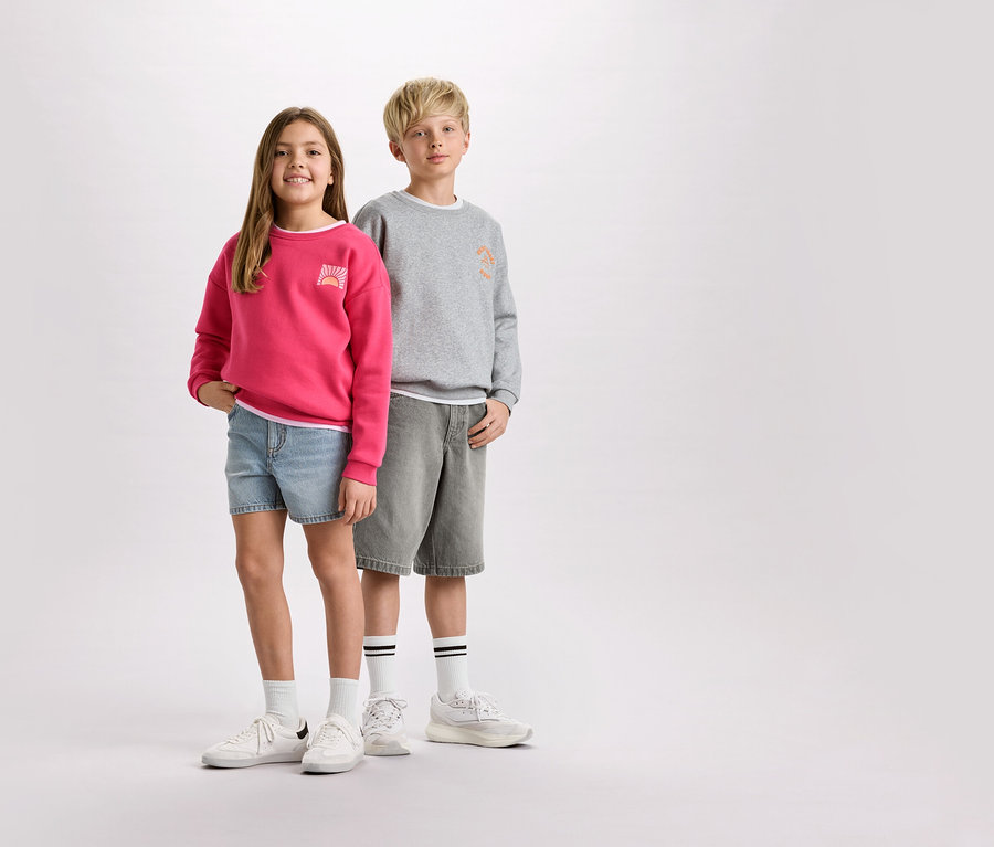 Une fille portant un pull rose et un short en jean se tient à côté d'un garçon portant un pull gris et un short.