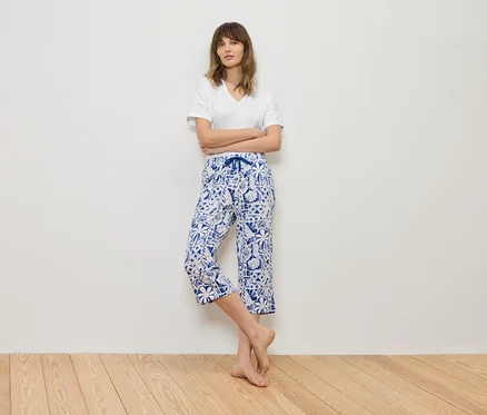 Femme debout, les bras croisés devant un mur, portant un t-shirt blanc et un pantacourt bleu à motifs.