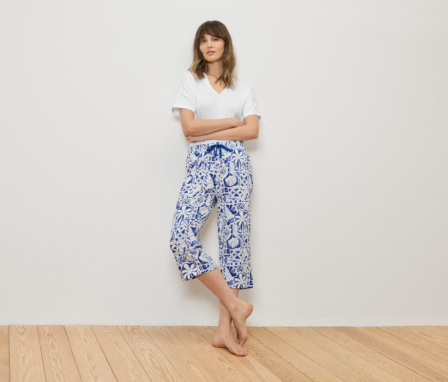 Femme debout, les bras croisés devant un mur, portant un t-shirt blanc et un pantacourt bleu à motifs.