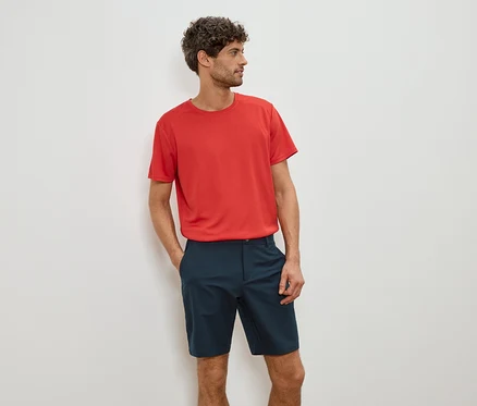 Homme portant un t-shirt rouge et un short bleu, les mains dans les poches.