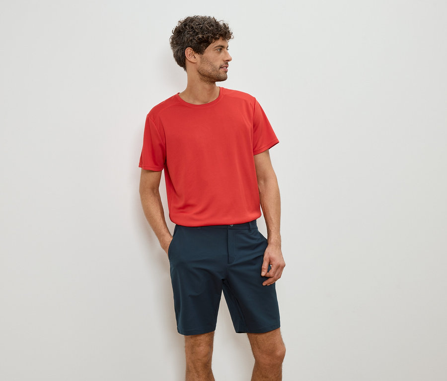 Homme portant un t-shirt rouge et un short bleu, les mains dans les poches.