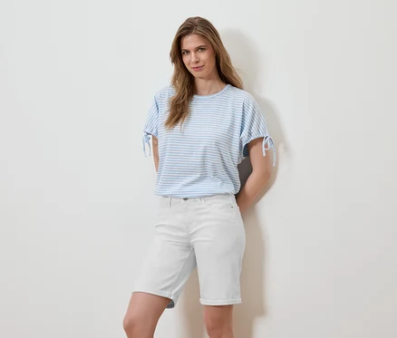 Une femme pose devant un mur blanc, portant un t-shirt rayé bleu clair et un short blanc.