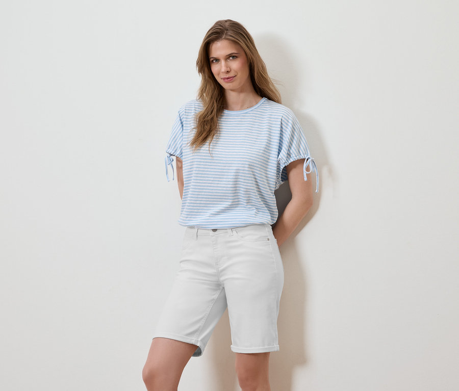 Une femme pose devant un mur blanc, portant un t-shirt rayé bleu clair et un short blanc.
