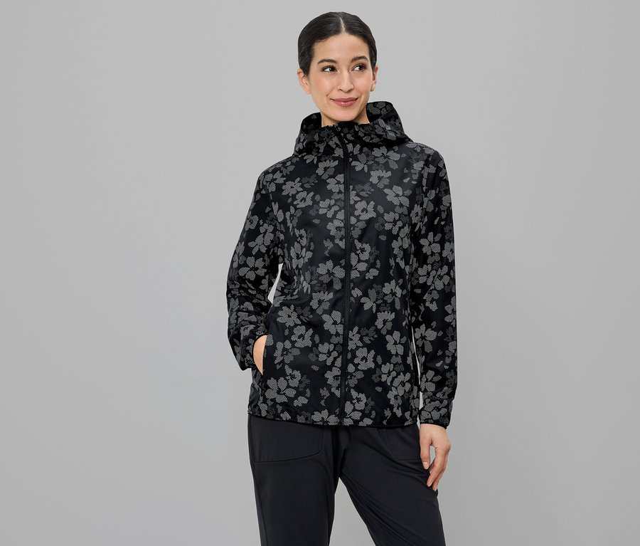 Une femme portant une veste à capuche à motif floral, la main dans la poche de son pantalon noir, sur un fond gris.