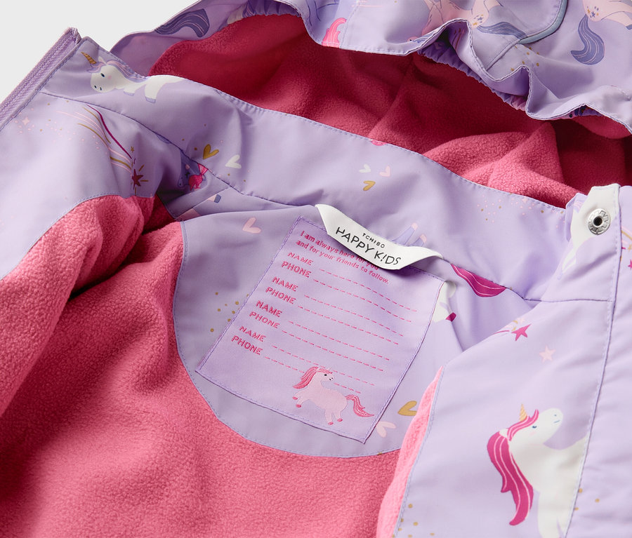 Gros plan d'une veste pour enfant violette à motif de licorne avec une doublure en polaire rose. Une étiquette avec "HAPPY KIDS" et un espace pour le nom/téléphone est visible.