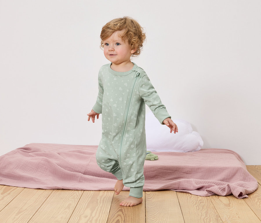 Un jeune enfant aux cheveux blonds bouclés marche pieds nus sur un plancher en bois. Il porte un pyjama vert clair avec une fermeture éclair et un motif de cœur.