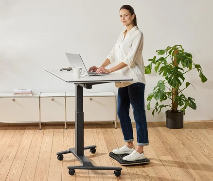 Une femme se tient debout à un bureau à hauteur réglable et utilise un ordinateur portable, une tasse est posée à côté.
